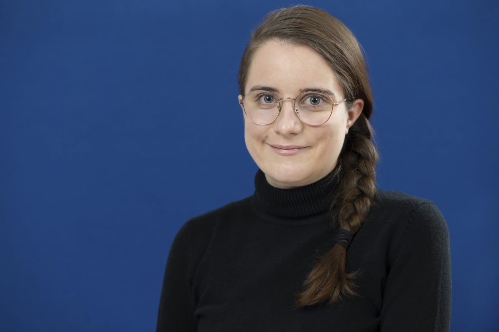 Eine Frau mit geflochtenem Zopf und Brille lächelt in die Kamera. Sie ist vor einem blauen Hintergrund.