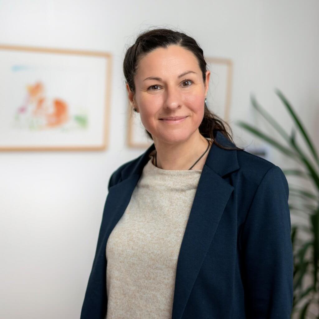 Eine Frau mit dunkelbraunen Haaren trägt einen blauen Blazer und lächelt in die Kamera.