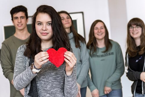 Eine junge Frau zeigt ein rotes Herz. Im Hintergrund sind Schüler:innen zu sehen, die sich freuen.