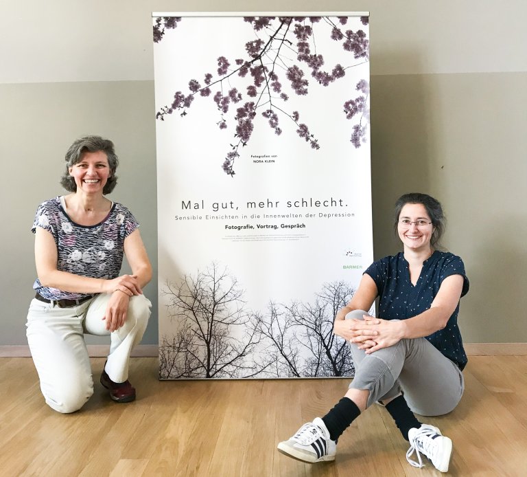 Zwei Frauen knien/sitzen neben einem Roll-Up auf dem steht: "Mal gut, mehr schlecht. Sensible Einsichten in die Innenwelten der Depression - Fotografie, Vortrag, Gespräch"