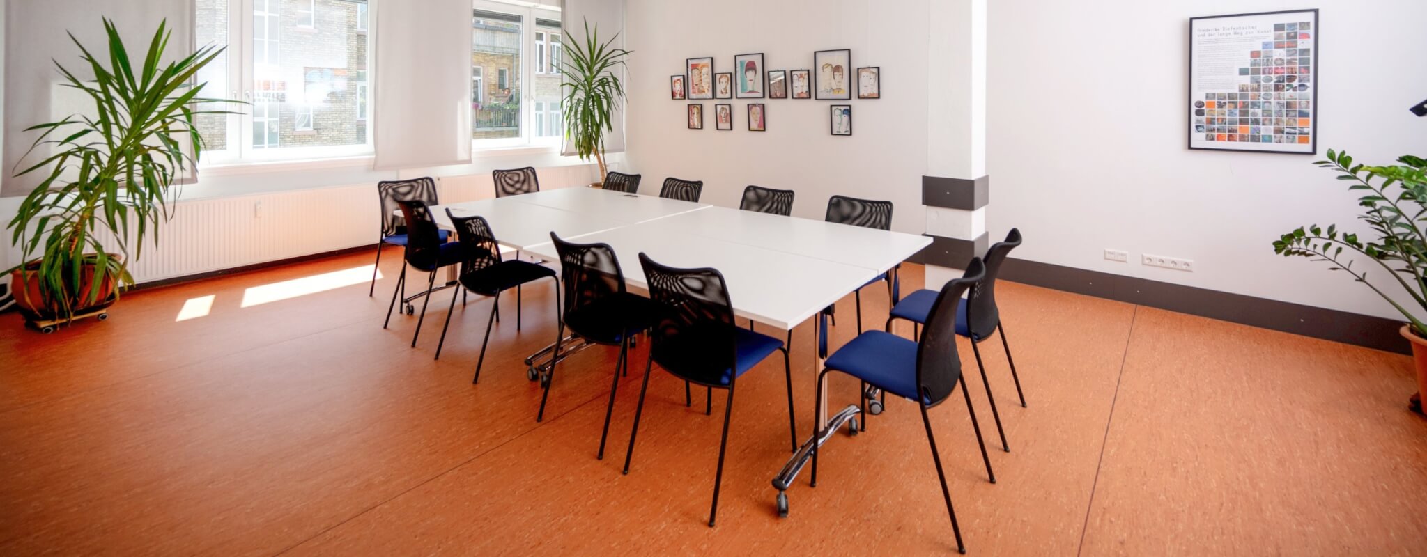 Zu sehen ist Raum 1.6: Tische mit 12 blauen Stühlen, Bilder von gezeichneten Köpfen an der Wand und 3 Pflanzen sin dzu sehen. Der Boden ist orange, es sind hellgraue Vorhänge zu sehen.